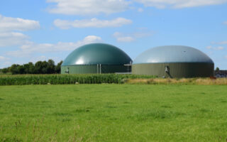 Photo of large green containments for anaerobic digestion. A green lawn is in the foreground and the structures are at a distance against a blue sky with clouds.